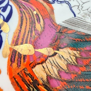 May include: Close-up of a decorative ceramic plate featuring a vibrant bird design. The bird has orange, pink, and teal feathers with gold leaf accents. Blue and gold decorative elements surround the bird on a white background.