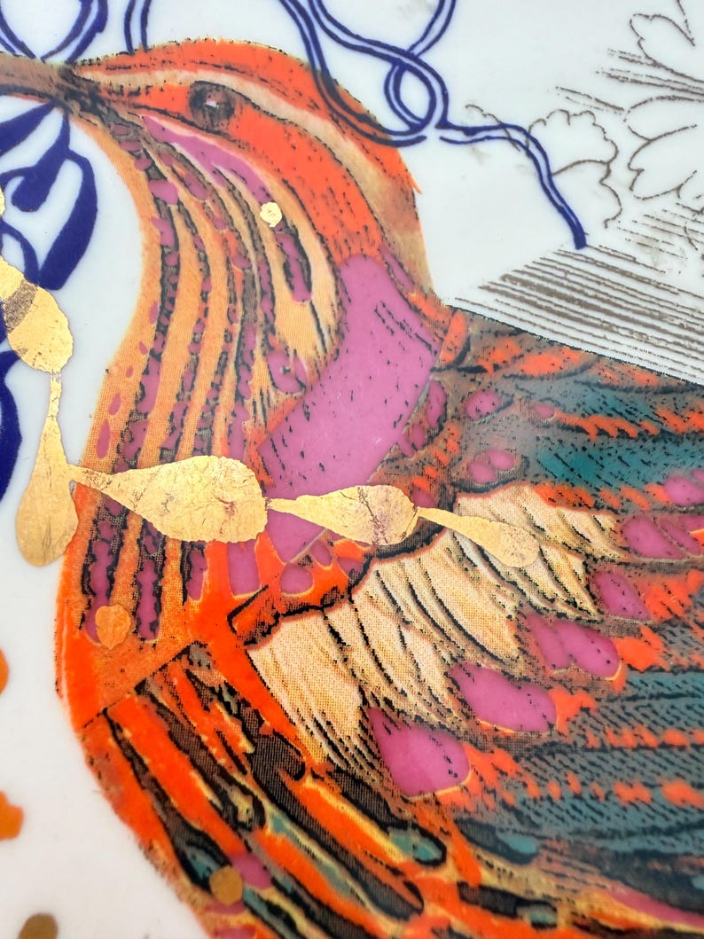 May include: Close-up of a decorative ceramic plate featuring a vibrant bird design. The bird has orange, pink, and teal feathers with gold leaf accents. Blue and gold decorative elements surround the bird on a white background.