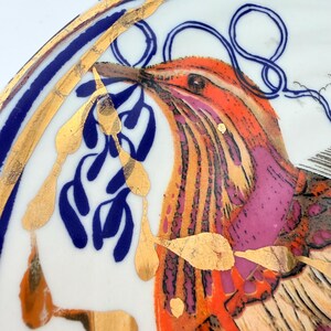 May include: A decorative plate with a bird design. The bird features orange, pink, and gold accents. The plate has a blue and gold border. The bird is holding blue leaves in its beak. The plate is white.