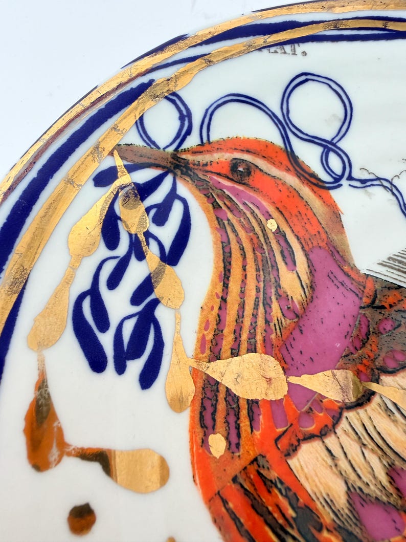 May include: A decorative plate with a bird design. The bird features orange, pink, and gold accents. The plate has a blue and gold border. The bird is holding blue leaves in its beak. The plate is white.