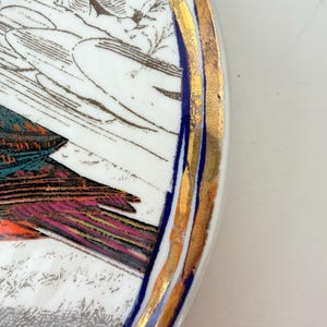 May include: A white ceramic plate with a detailed illustration of a bird and foliage. The plate has a gold rim with a blue accent. The bird's feathers are shades of teal, pink, and orange.