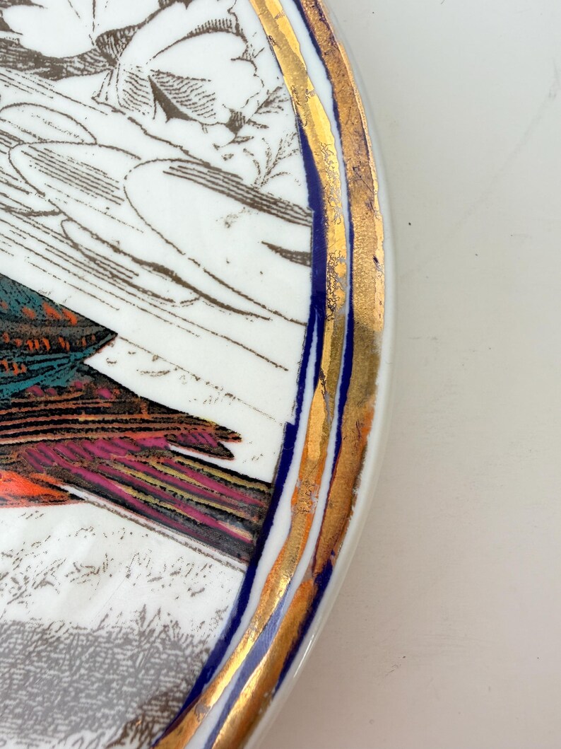 May include: A white ceramic plate with a detailed illustration of a bird and foliage. The plate has a gold rim with a blue accent. The bird's feathers are shades of teal, pink, and orange.