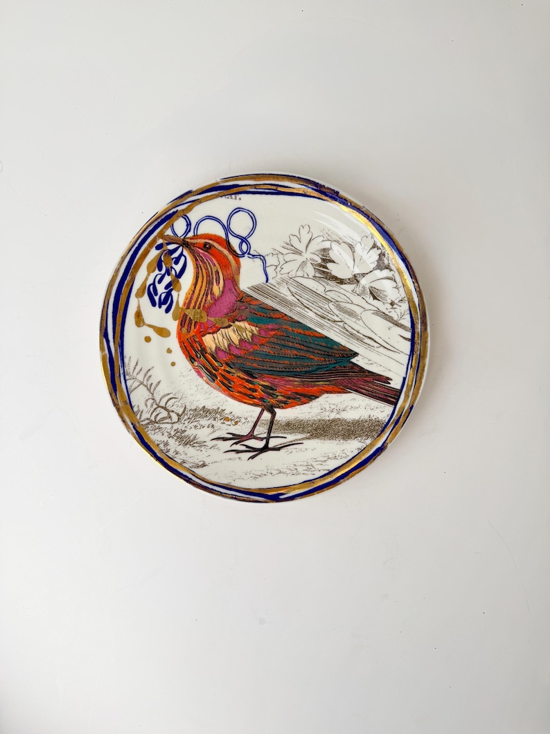 May include: A decorative plate featuring a colorful bird illustration. The bird has red, orange, and purple feathers, and is holding a blue sprig. The plate has a gold rim, a blue border, and a white background with floral and gold accents.
