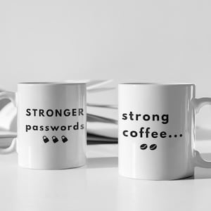 Puede incluir: Dos tazas de cerámica blancas con texto y diseños negros. Una taza dice "STRONGER passwords!" con tres iconos de candado. La otra dice "strong coffee..." con iconos de granos de café. Una pequeña planta en una maceta blanca está en el fondo.