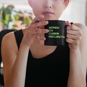 May include: A black mug with the text "WOMEN IN CYBER SECURITY" in green, held by a person wearing a black vest top. The mug is the central object, with the text clearly displayed. The background is out of focus.