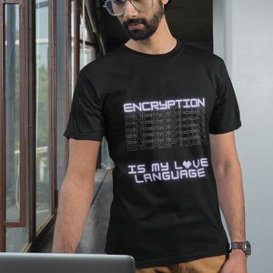 May include: Black t-shirt with the text "ENCRYPTION IS MY LOVE LANGUAGE" in a white, futuristic font. The shirt is worn by a person with glasses and a beard, standing near a window and a laptop.