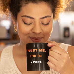 May include: A black coffee mug with the text "TRUST ME. I'M IN /root" in orange. The mug is held by a person wearing a white top. The person has curly brown hair and is looking down at the mug.