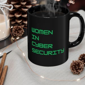 May include: A black ceramic mug with the text "WOMEN IN CYBER SECURITY" in green. The mug contains a dark liquid, with steam rising. It sits on a light grey surface, alongside decorative elements, including a candle and pine cones.
