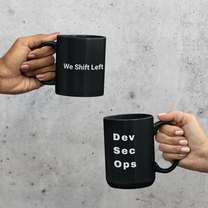May include: Two black coffee mugs with white text are held up against a grey background. One mug reads "We Shift Left," and the other reads "Dev Sec Ops." The mugs have a simple, modern design.