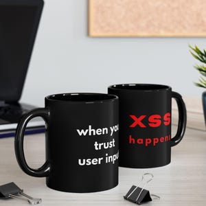 May include: Two black ceramic mugs with white and red text. One mug reads "when you trust user input" in white. The other mug reads "XSS happens" in red. The mugs are on a light-coloured surface.