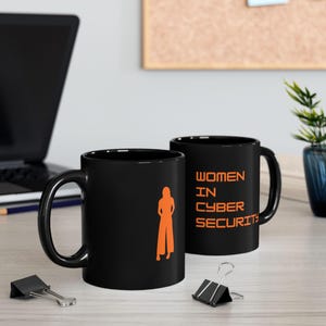 May include: Two black coffee mugs with orange graphics. One mug features a silhouette of a person, the other mug has the text "WOMEN IN CYBER SECURITY". The mugs are on a light wood surface with a laptop and office supplies in the background.