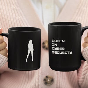 May include: Two black coffee mugs. One mug has a white silhouette of a woman. The other mug has the text "WOMEN IN CYBER SECURITY" in white. The mugs are held by a person wearing a pink cardigan.
