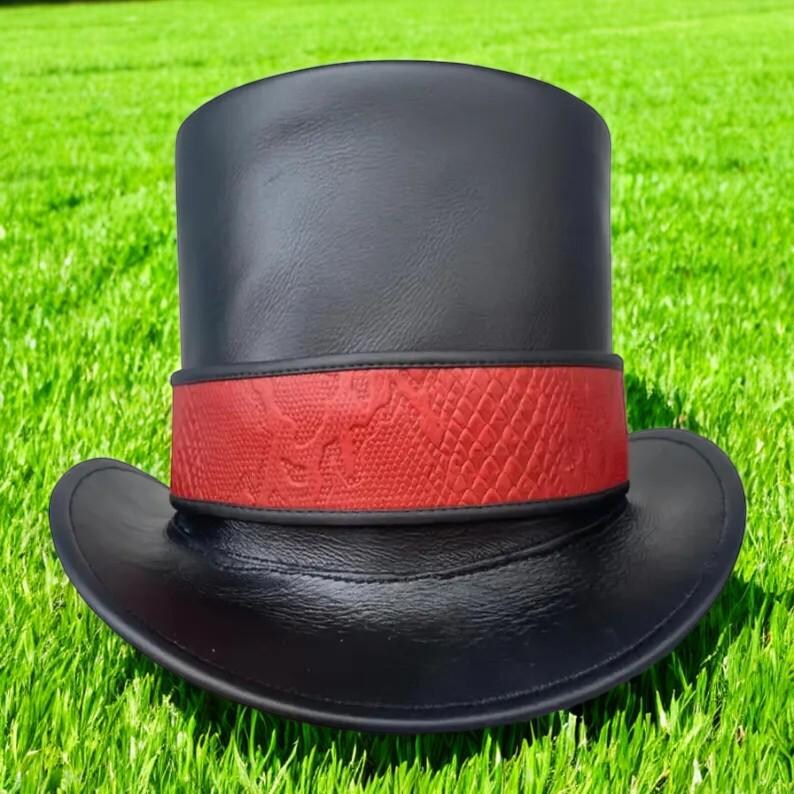 Op de afbeelding: Een zwarte hoge hoed met een rode band met slangenpatroon. De hoed is gemaakt van een glad, glanzend materiaal en ligt op een groene grasmat. Hij heeft een brede rand en een hoge, cilindrische kroon.