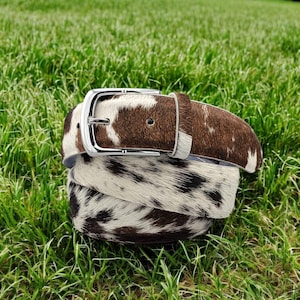 May include: A rolled belt made from cowhide, featuring a brown and white spotted pattern. It has a silver-coloured buckle. The belt is displayed on green grass. This is a fashion accessory.