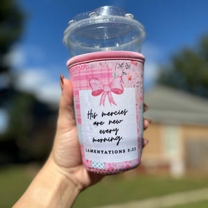 May include: A pink and white coffee cup sleeve with a bow and floral design. The sleeve has the text "His mercies are new every morning. Lamentations 3:23". A clear plastic cup with a lid is on top.