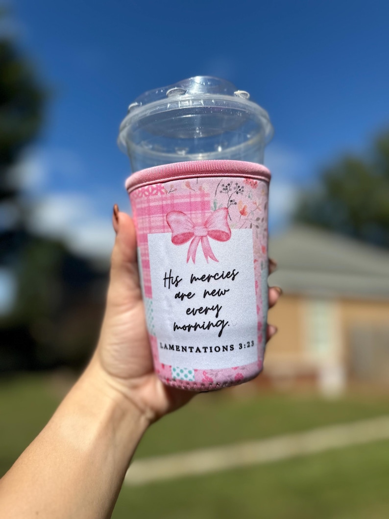 May include: A pink and white coffee cup sleeve with a bow and floral design. The sleeve has the text "His mercies are new every morning. Lamentations 3:23". A clear plastic cup with a lid is on top.