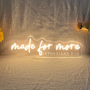 Könnte beinhalten: Ein Neonschild mit dem Schriftzug "made for more" in Schreibschrift. Darunter steht "Ephesians 2:10". Das Schild leuchtet in einem warmen, sanften Licht. Eine Sonnenblume und orangefarbene Blumen sind im Hintergrund.