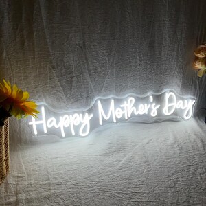 May include: A white neon sign that reads "Happy Mother's Day" in a cursive font. The sign is illuminated and appears to be made of flexible tubing. The background is a white fabric. A bouquet of yellow flowers is visible on the left.