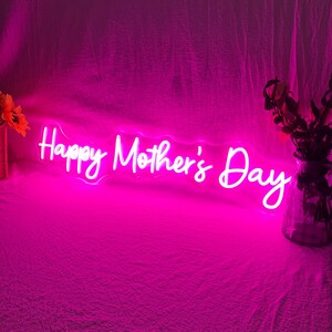 May include: A neon sign in bright pink displays the words "Happy Mother's Day" in a cursive font. The sign is illuminated against a pink backdrop, with floral arrangements on either side.