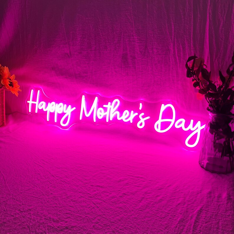 May include: A neon sign in bright pink displays the words "Happy Mother's Day" in a cursive font. The sign is illuminated against a pink backdrop, with floral arrangements on either side.