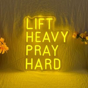 May include: A neon sign with the words "LIFT HEAVY PRAY HARD" in bright yellow. The sign is against a yellow background with some flowers in the corners. The sign is a motivational piece.