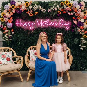 May include: A woman and a young girl pose in front of a floral backdrop with a neon sign that reads "Happy Mother's Day." The woman wears a blue dress, and the girl wears a pink dress. Two wicker chairs with floral pillows are in the foreground.