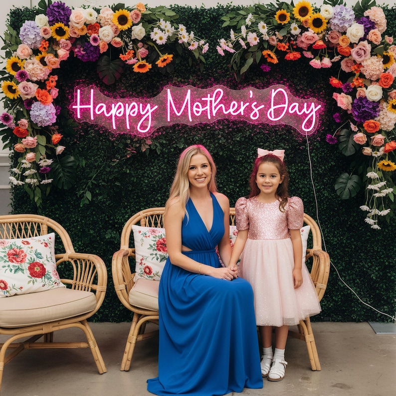 May include: A woman and a young girl pose in front of a floral backdrop with a neon sign that reads "Happy Mother's Day." The woman wears a blue dress, and the girl wears a pink dress. Two wicker chairs with floral pillows are in the foreground.