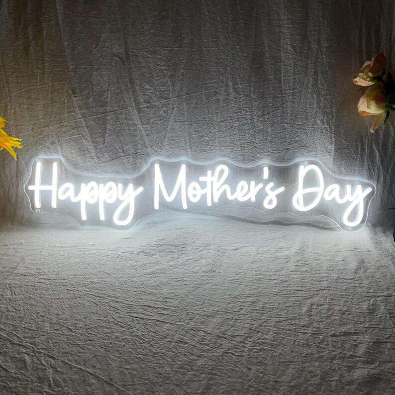 May include: A white neon sign that reads "Happy Mother's Day" in a cursive font. The sign is illuminated and appears to be made of flexible tubing. The background is a textured, off-white fabric.