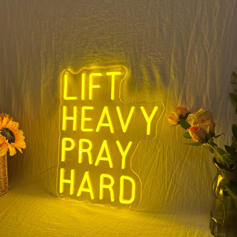May include: A bright yellow neon sign with the words "LIFT HEAVY PRAY HARD" in a bold, sans-serif font. The sign is illuminated against a yellow backdrop, with a sunflower and flowers in a vase on either side.