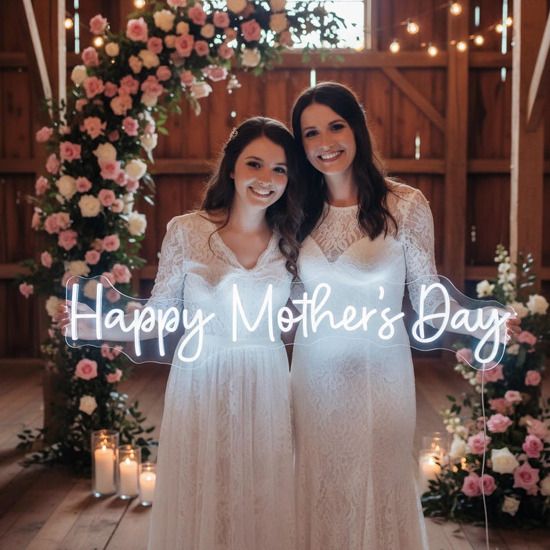 May include: Two people in white lace dresses smile, holding a neon sign that reads "Happy Mother's Day." The background features a floral arch of pink and white roses, candles, and wooden beams, creating a warm, celebratory atmosphere.
