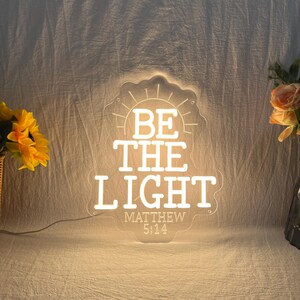 May include: A clear acrylic LED sign with a scalloped edge, illuminated with warm white light. The text reads BE THE LIGHT followed by MATTHEW 5:14. A sunburst graphic sits above the text. The sign is powered by a thin cord extending from the base.