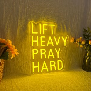 May include: A yellow neon sign with the words "LIFT HEAVY PRAY HARD" in a stacked layout. The sign is illuminated against a yellow background. Decorative flowers are visible on either side of the sign.