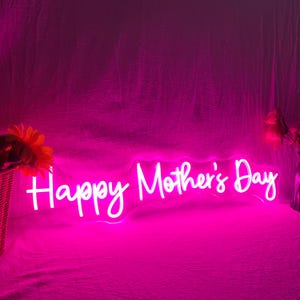 May include: A neon sign in bright pink illuminates the words "Happy Mother's Day". The sign is in a cursive font and is set against a pink backdrop. A basket of flowers is visible on the left.