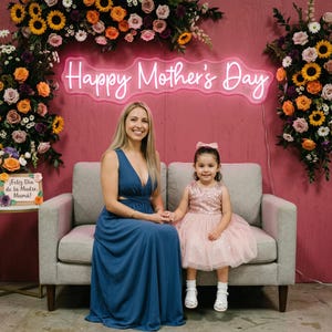 May include: A woman and a young girl sit on a gray couch in front of a pink wall decorated with flowers. A neon sign reads "Happy Mother's Day." The woman wears a blue dress, and the girl wears a pink dress. A sign on the left says "Feliz D&iacute;a de la Madre, Mam&aacute;!"