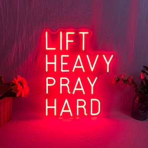 May include: A bright red neon sign with the words "LIFT HEAVY PRAY HARD" in a stacked layout. The sign is illuminated against a soft, pinkish background, with a basket of flowers and a vase of flowers in the corners.