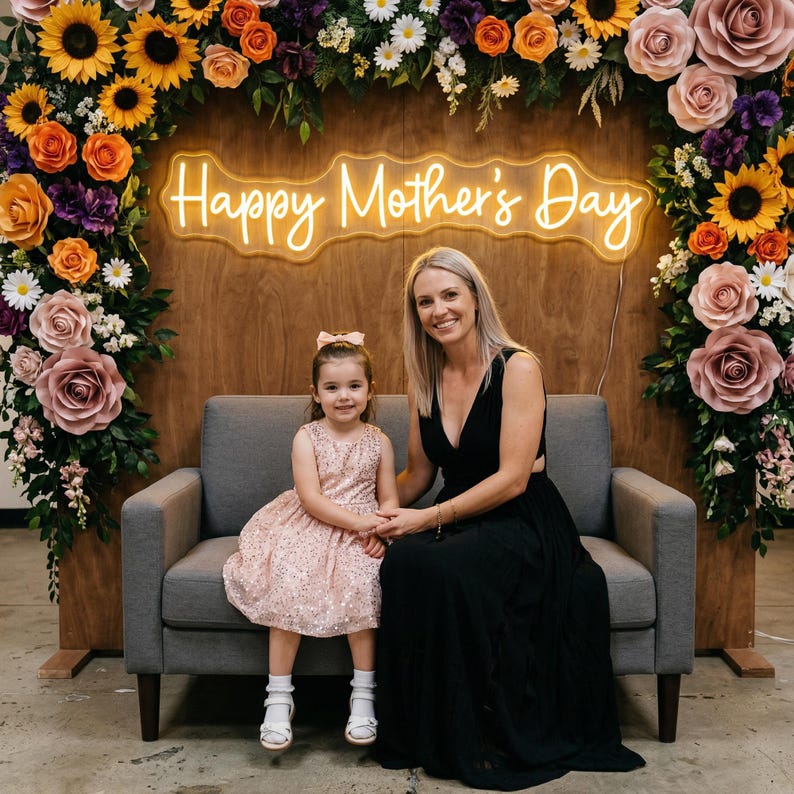 May include: A woman and a young girl sit on a gray couch in front of a wooden backdrop. The backdrop features a neon sign that reads "Happy Mother's Day" surrounded by floral arrangements of sunflowers, roses, and daisies. The girl wears a pink dress.
