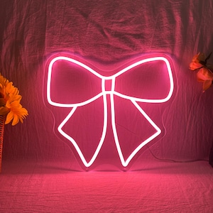 Può includere: Un'insegna al neon a forma di fiocco, che emette una luce rosa brillante. Il cartello è delineato in bianco e si trova su uno sfondo rosa. Il design del fiocco è simmetrico, con due anelli e due nastri.