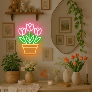 May include: A neon sign in the shape of tulips in a pot, with pink flowers, green leaves, and an orange pot. The sign is mounted on a wall with framed artwork, a mirror, and a shelf with decorative items.
