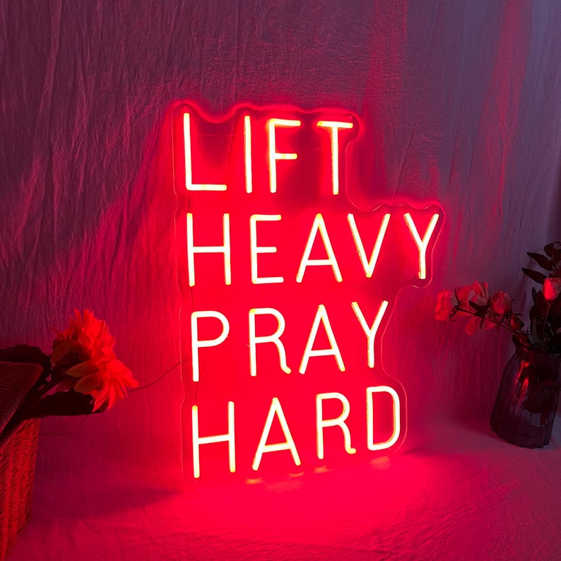 May include: A red neon sign with the words "LIFT HEAVY PRAY HARD" in a bold, sans-serif font. The sign is illuminated against a pink background, with flowers in a vase and a basket in the corners.