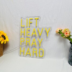 May include: A clear acrylic sign with the words "LIFT HEAVY PRAY HARD" in bright yellow neon. The sign is shaped like a cloud. A sunflower and a vase of flowers are in the background.