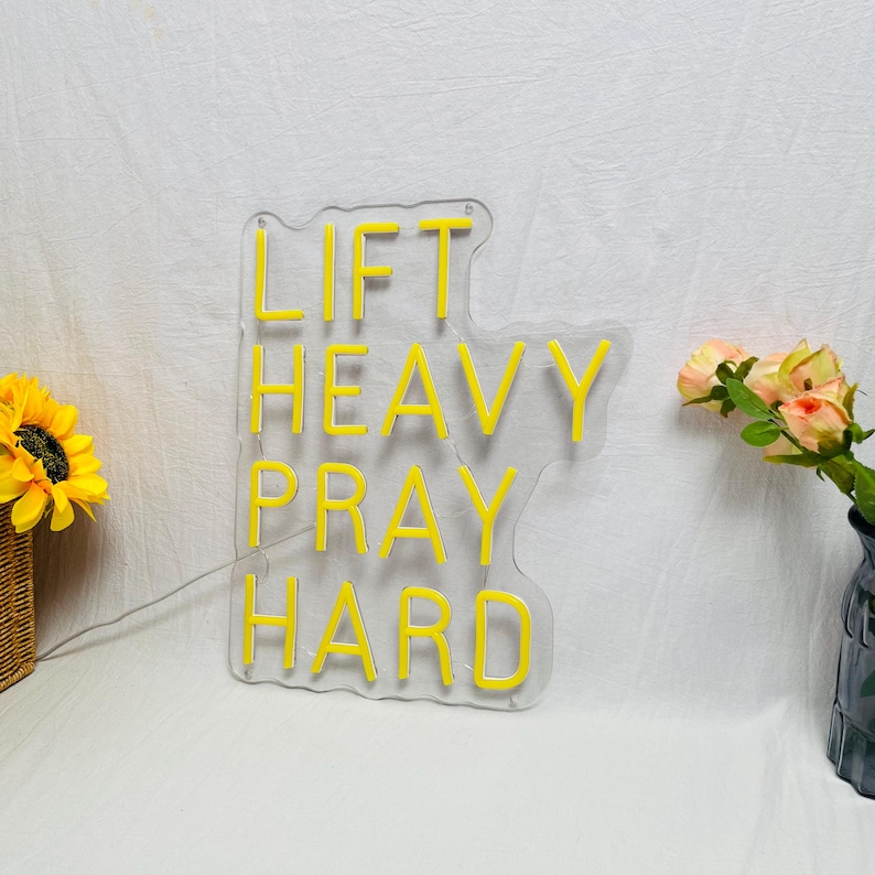 May include: A clear acrylic sign with the words "LIFT HEAVY PRAY HARD" in bright yellow neon. The sign is shaped like a cloud. A sunflower and a vase of flowers are in the background.