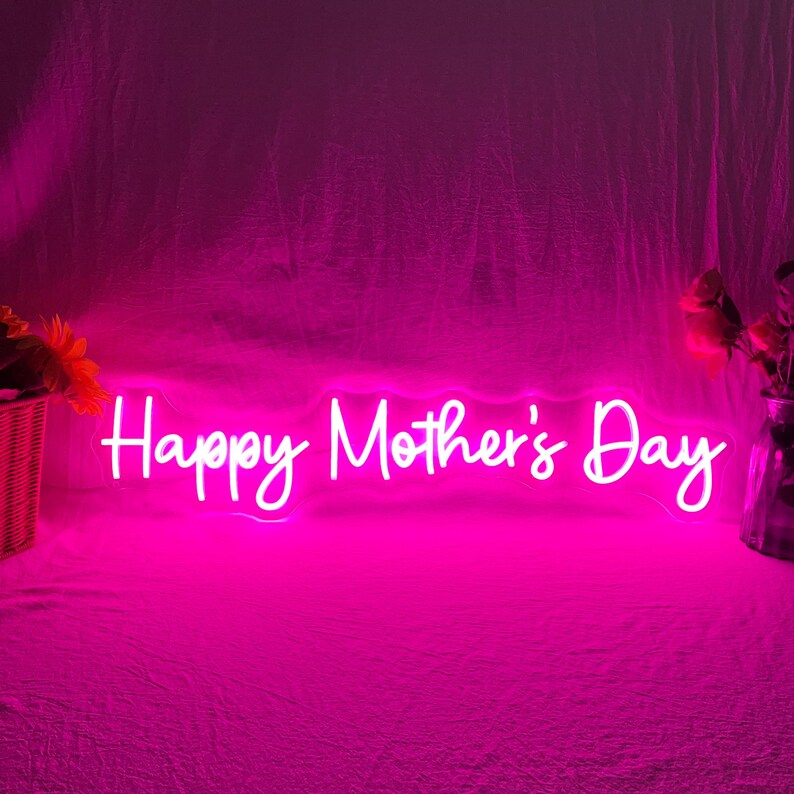 May include: A neon sign in bright pink displays the words "Happy Mother's Day" in a cursive font. The sign is illuminated against a pink backdrop, with a basket of flowers on the left and a vase of flowers on the right.