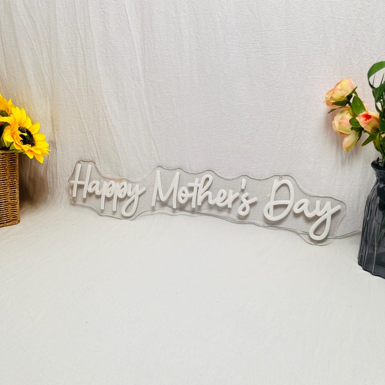 May include: A white neon sign with the words "Happy Mother's Day" in a cursive font. The sign is on a clear backing. A woven basket with sunflowers and a vase with flowers are on either side of the sign.