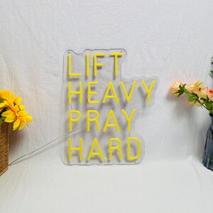 May include: A clear acrylic sign with the words "LIFT HEAVY PRAY HARD" in bright yellow neon. The sign is on a white surface, with a small bouquet of flowers to the side.