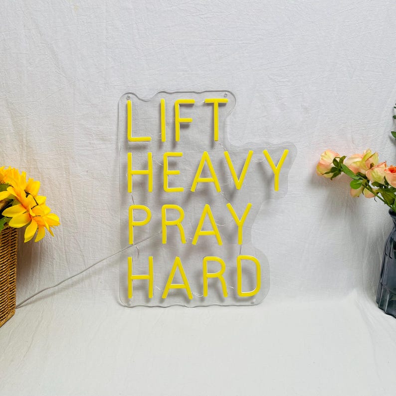 May include: A clear acrylic sign with the words "LIFT HEAVY PRAY HARD" in bright yellow neon. The sign is on a white surface, with a small bouquet of flowers to the side.