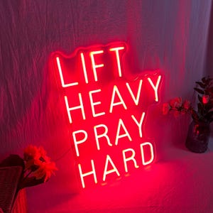 May include: A red neon sign with the words "LIFT HEAVY PRAY HARD" in white lettering. The sign is illuminated and has a decorative shape. The background is a soft, pinkish-red fabric.