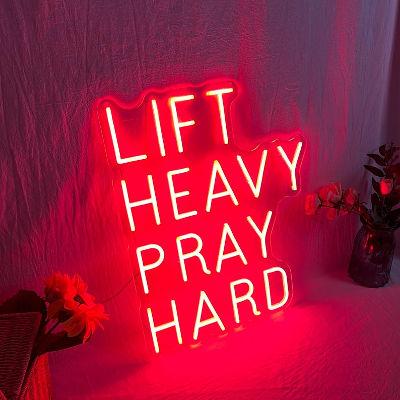 May include: A red neon sign with the words "LIFT HEAVY PRAY HARD" in white lettering. The sign is illuminated and has a decorative shape. The background is a soft, pinkish-red fabric.
