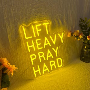 May include: A yellow neon sign with the words "LIFT HEAVY PRAY HARD" in a stacked layout. The sign is illuminated against a yellow backdrop, with a vase of flowers to the right and a bouquet of flowers to the left.