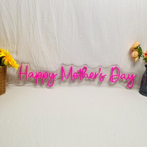 May include: A neon sign in a cursive font that reads "Happy Mother's Day" in bright pink. The sign is mounted on a clear, wavy-edged backing. The background is a white fabric with a basket of yellow flowers and a vase of peach-colored roses on either side.