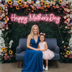 May include: A woman and a young girl sit on a blue couch in front of a floral backdrop. The woman wears a long, blue dress, and the girl wears a pink dress. A neon sign above them reads "Happy Mother's Day."